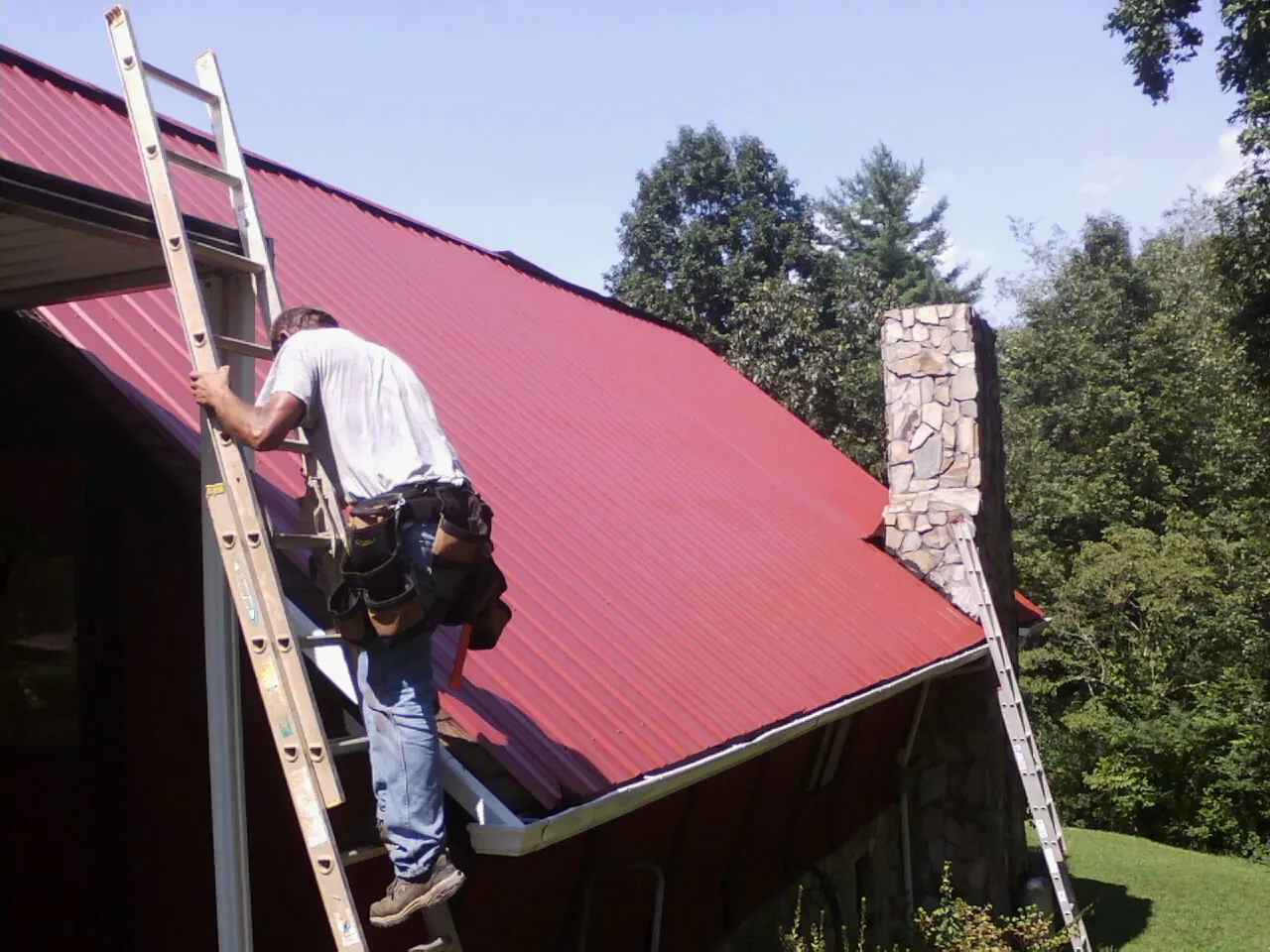 Roofer on a ladder beside a red metal roof and stone chimney — Soffit & Fascia Repair by Roofing Brothers in La Valle